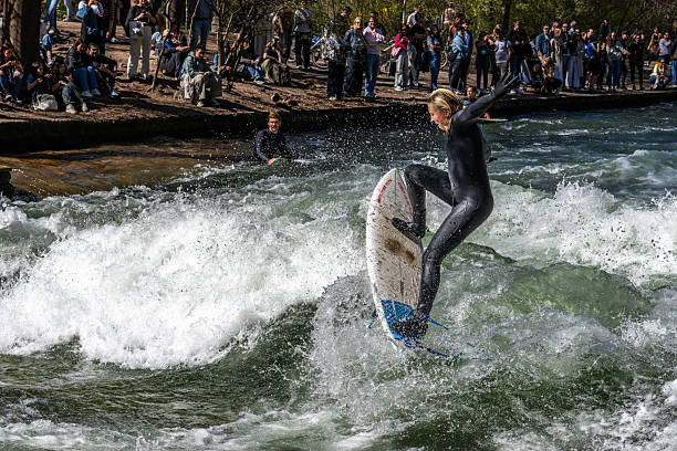 Munich, Germany - Apr 12, 2025: Surfer in the city river, Munich is famous for people surfing in urban enviroment called Eisbach. Surfers practicing their skill on an artificial wave in central Munich