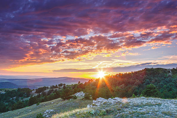 A breathtaking sunset illuminates a mountain landscape, casting vibrant orange and purple hues across the clouds. The high-angle view captures the serene beauty of rolling green hills and forests under a dramatic sky, evoking a feeling of tranquility and majesty.