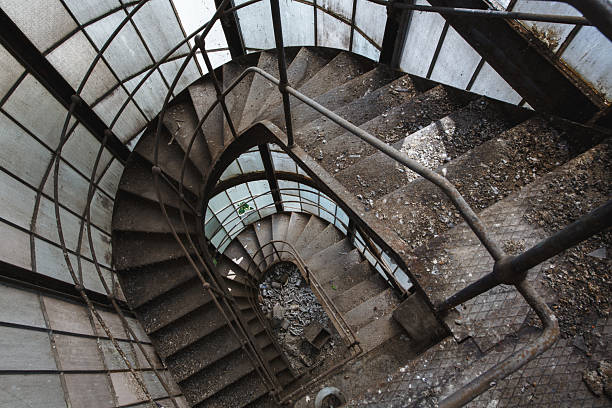 This evocative image captures a rusted metal spiral staircase winding down through a derelict industrial tower, surrounded by glass grid windows. The scene conveys a powerful sense of urban decay and forgotten history, perfect for themes of isolation, neglect, and architectural exploration.