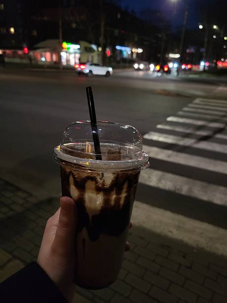 Hand holding iced coffee cup on a city street at night with blurred urban background and pedestrian crossing