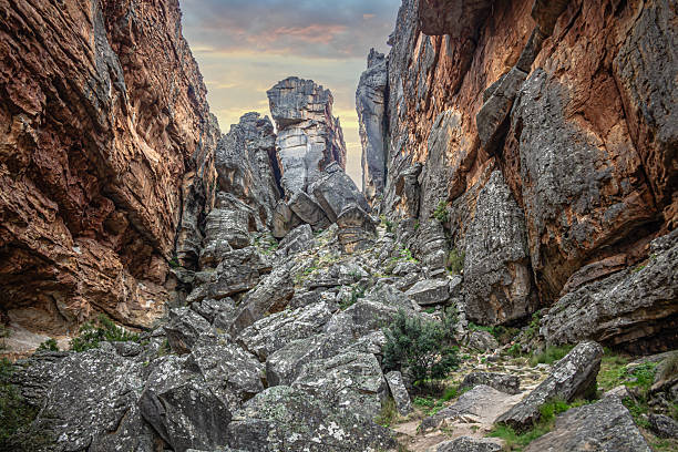 Dramatic rocky canyon scene with towering weathered sandstone cliffs, massive boulders, and rugged terrain under a cloudy sky at dusk. Remote wilderness landscape showcasing geological formations, natural textures, and the raw beauty of an untamed environment.