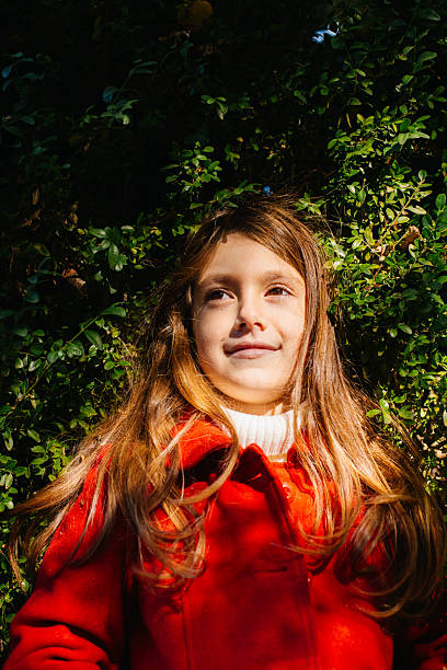 Portrait of a young girl wearing a red coat, smiling gently while standing outdoors against dense green foliage. Warm sunlight highlights her face, creating a natural lifestyle scene in a garden or park setting.