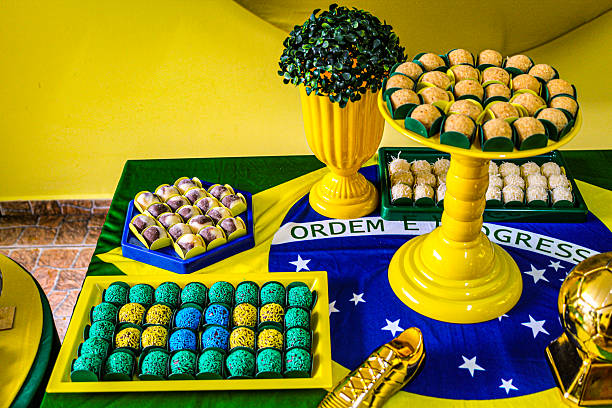 A beautifully decorated Brazilian dessert table featuring a variety of colorful sweets and treats, including brigadeiros, chocolates, and truffles, all arranged on a vibrant yellow, green, and blue tablecloth with a Brazilian flag design, complete with a decorative tree and vase, and a joyful atmosphere perfect for a festive celebration or party.