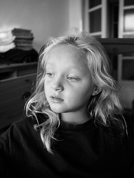 6-year-old girl wearing a translucent head covering smiles gently while touching her face with both hands. The black and white film photograph captures her soft expression in close-up. Light falls across her features from a nearby window, creating a dreamy, nostalgic mood. The grain and contrast of analog film photography adds texture and depth to this vintage-style portrait.