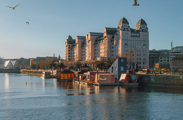 Oslo, Norway - Winter: Havnelageret, also known as the Pink Palace in Bjørvika, seen along the Oslo harbor at Langkaia, illuminated by warm winter sunlight and reflected on calm blue water. Originally completed in 1921 as one of Europe’s largest concrete buildings, the historic waterfront structure now serves as a modern commercial office building, capturing the blend of industrial heritage and contemporary urban development in Oslo’s harbor district