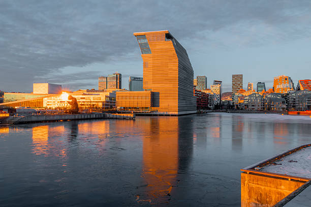 Panoramic view of modern waterfront architecture and contemporary urban skyline along the Oslo harbor area in Norway on a sunny winter day at sunset. Warm golden hour light illuminates glass and metal buildings, creating clear reflections on the calm fjord water. The scene highlights Scandinavian urban renewal, modern city life, sustainable architecture, and winter atmosphere in a redeveloped waterfront district built on a former industrial harbor