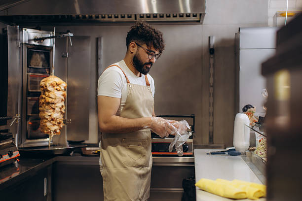 Professional chef wearing an apron and gloves, preparing shawarma meat on a vertical rotisserie grill in a clean commercial kitchen