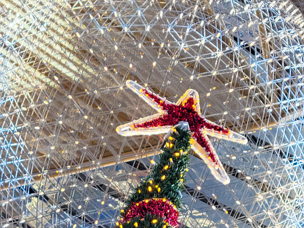 A creative holiday decoration features a translucent star with embedded red lights serving as the topper for a small artificial Christmas tree wrapped in green tinsel and red ornaments. The tree stands against a backdrop of a gleaming metallic grid structure, suggesting an indoor architectural space like a contemporary atrium or dome. No people are present. The image captures a whimsical fusion of marine and festive elements, with warm red and green glows contrasting the cool silver tones of the surroundings. Ideal for commercial uses in holiday marketing campaigns, interior design inspirations for public spaces, marine-themed seasonal promotions, or editorial content on innovative decorations during winter festivities.
