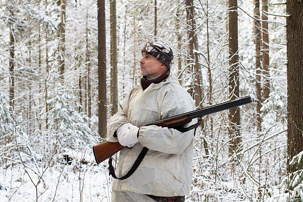 A hunter dressed in winter camouflage with a shotgun in his hands stands in a snowy winter forest.