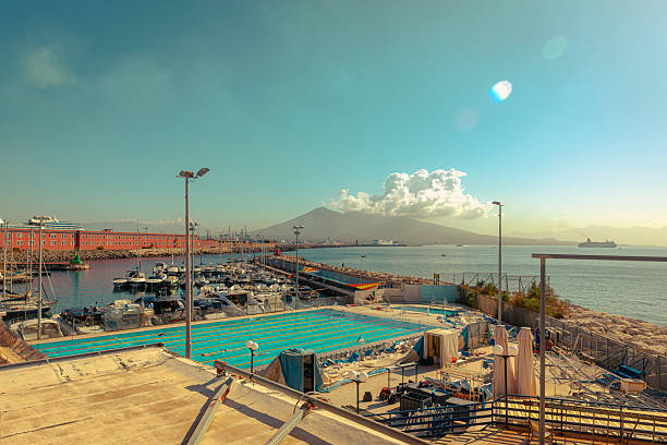 Wide panoramic view of the Naples waterfront featuring a marina, coastal swimming facilities, and urban seaside infrastructure, with Mount Vesuvius rising in the background under a clear sky. The scene captures the blend of maritime life, city architecture, and natural landscape that defines Naples and the Gulf of Naples, conveying a calm yet vibrant Mediterranean atmosphere.