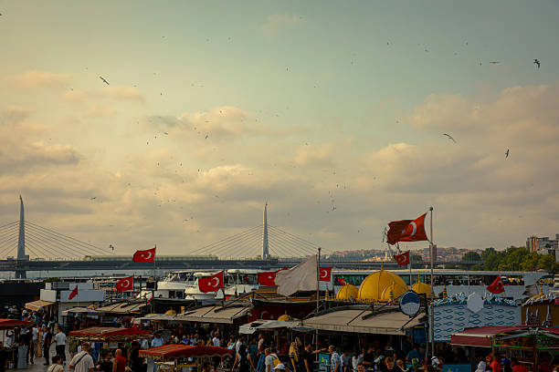 Vibrant evening scene at the Eminönü waterfront in Istanbul, with food stalls, flags, boats, and crowds of locals and tourists. In the background stands the modern Golden Horn Metro Bridge, while seagulls fill the warm sky above the harbor. The lively atmosphere captures the unique blend of tradition, commerce, and daily life that defines this bustling area of the city.