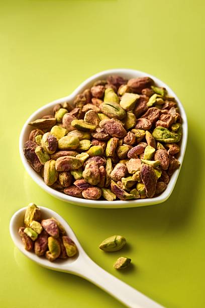 Pistachio nuts without shell in a heart shaped bowl on a green background. Spoon with pistachios. Green color. High angle view