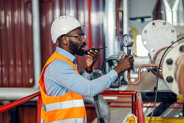 African american engineer in hardhat and high-visibility vest inspects a pressure gauge and speaks into a walkie-talkie in a boiler room, focused and professional