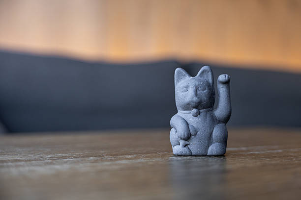 A black ceramic cat figurine with a raised paw sits on a wooden table. The background is softly blurred, creating a warm atmosphere.