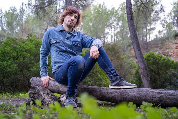 Young adult man in a denim jacket and jeans sitting casually on a fallen tree trunk in a tranquil forest setting, finding a moment of solitude and connection with nature