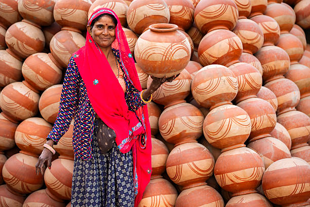 A smiling woman in bright traditional Rajasthani clothing and a red dupatta holds a decorated terracotta pot against a backdrop of neatly stacked earthen pots. The image highlights vivid colors, handmade pottery patterns, and a lively market or craft setting.