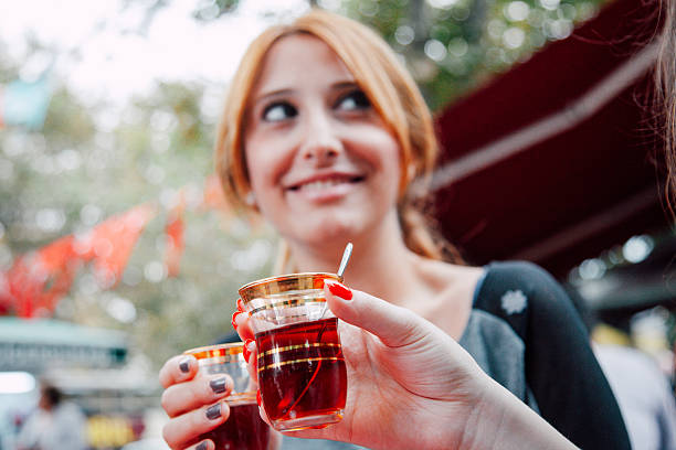 Young woman smiling and looking away while holding a glass of Turkish tea in an outdoor café environment, capturing a relaxed and cheerful lifestyle moment.