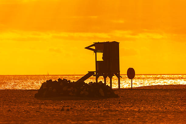 Lifeguard tower is silhouetted against a vibrant orange sunset on a sandy beach. A pile of rocks and a red ring sit in the foreground near the ocean in the twilight