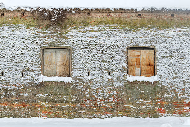 Old brick fortress wall with two windows partially covered in snow, showing weathered textures and historical architecture in winter.