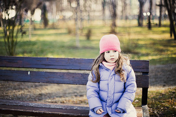 Color photograph of a young child sitting quietly on a wooden bench in a park during a cool season. Peaceful lifestyle scene capturing childhood emotions, contemplation, calmness, and everyday outdoor life in a natural setting.