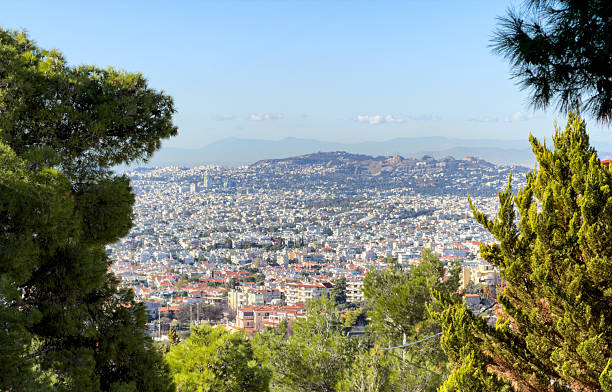 Athens cityscape from Lycabettus Hill framed by pine trees, looking over dense rooftops, distant mountains, calm blue sky.