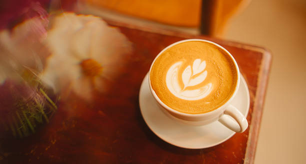 Latte coffee cup with leaf latte art on wooden table with blurred flower in warm light, cozy atmosphere for relaxing moment