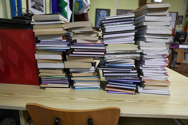 Large Stack Of Books And Notebooks On A Wooden Desk In Classroom Or Office Interior, Representing Education, Knowledge, Studying, Research, Workload, Learning Process, And Academic Pressure Concept.