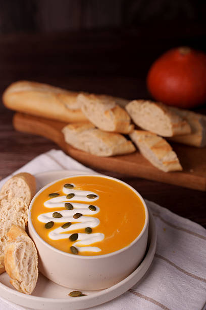 warm pumpkin soup garnished with cream and seeds served with freshly sliced baguette on a wooden board and white cloth with autumn pumpkin background, photograph, closeup, view, arrangement