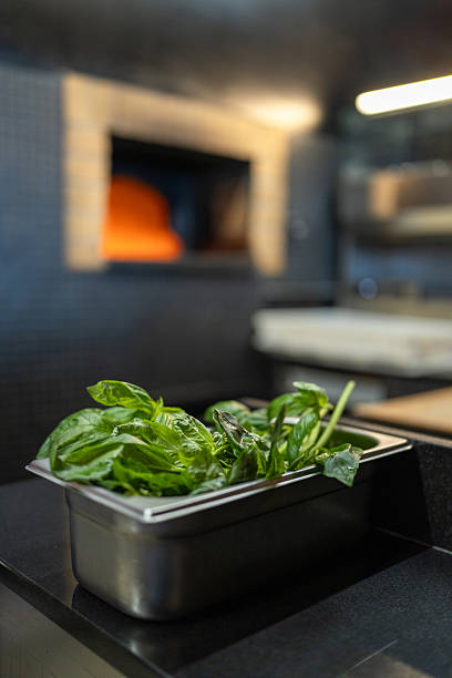 Fresh green salad in metal container resting on dark countertop in professional restaurant kitchen. Blurred background showing hot pizza oven with orange glow, ready for cooking.