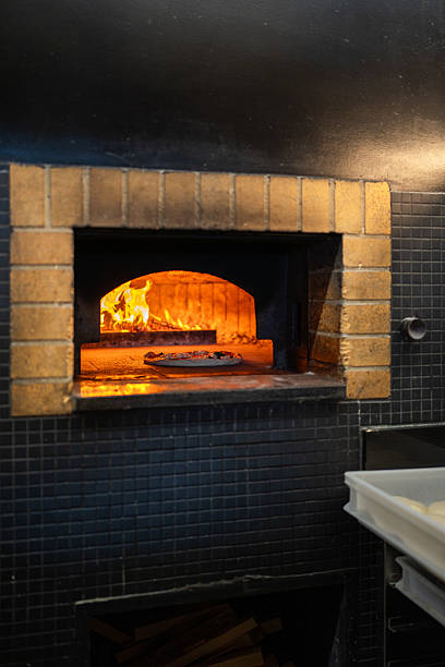 Warm vibrant flames burning inside a traditional stone oven, framed by brickwork. An uncooked pizza baking on the hearth, slowly cooking with intense heat. This commercial kitchen appliance preparing food for future restaurant customers.