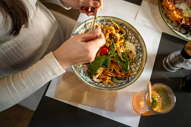 Hands of a person enjoying a delicious Italian pasta dish, dining at a restaurant table. The close-up shot focuses on the meal, with fresh ingredients and chopsticks being used to eat the noodles.