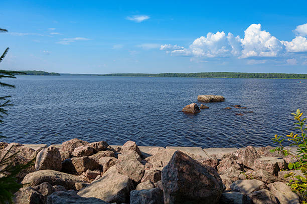 Wide view of a calm lake with a rocky shoreline in the foreground and scattered stones in the water, under a bright blue sky with clouds on a summer day