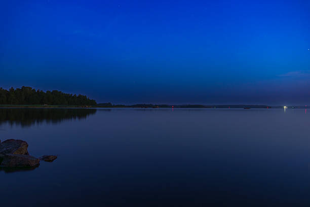 Peaceful lake scene at twilight with still water, scattered rocks in the foreground, and distant lights from boats on the horizon under a deep blue sky