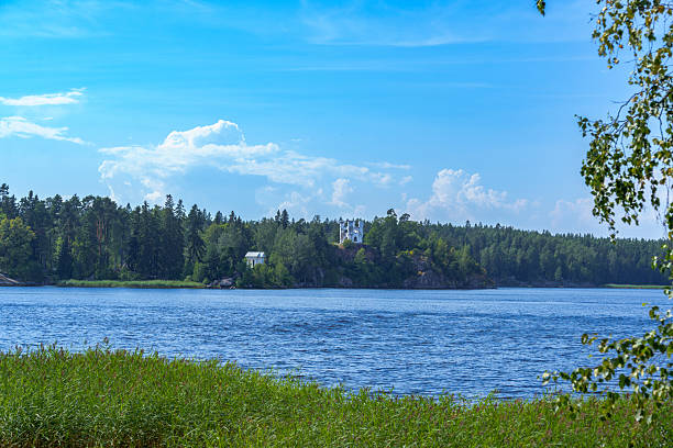 A white historic castle or tower stands atop a rocky, forested hill overlooking a calm lake, surrounded by dense greenery under a bright summer sky
