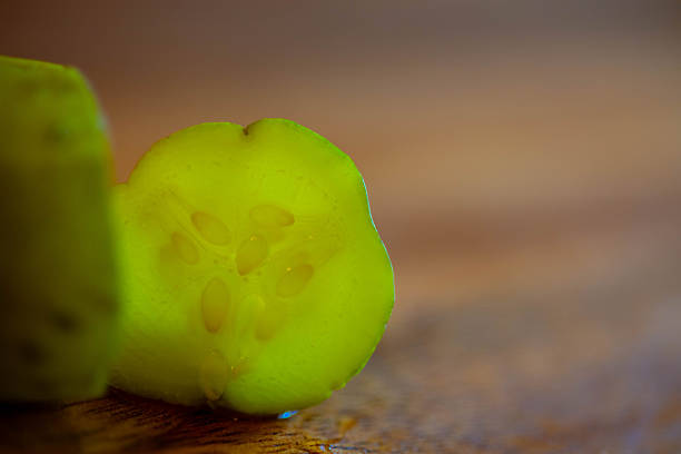 A close-up, backlit macro shot of a single slice of pickled cucumber standing vertically on a wooden cutting board. The strong backlighting shines through the translucent green flesh, highlighting the internal seed structure and texture against a soft, dark blurred background.