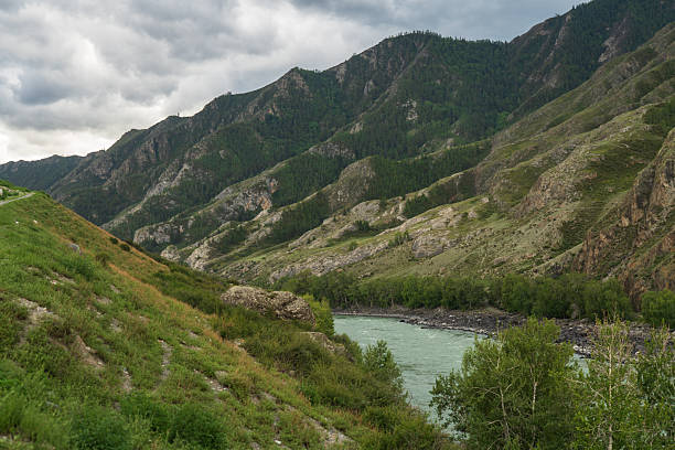Wide view of a mountain river flowing through a green valley with steep hills under cloudy sky. Suitable for nature, environment, and wilderness background use.