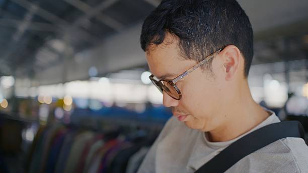 Conscious Asian man carefully choosing second-hand clothing at a thrift shop. This image illustrates the growing trend of sustainable fashion, emphasizing the benefits of saving money, reducing waste, and conserving global resources through circular consumption.