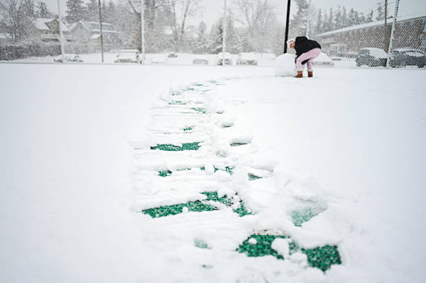 Child enjoying winter by rolling a large snowball across a snow-covered sports field