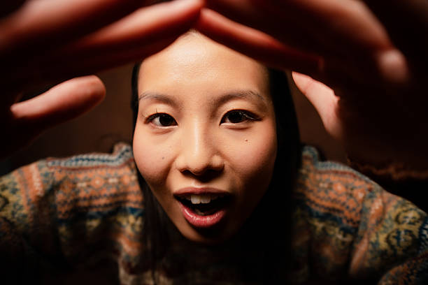 Asian young adult woman making a playful gesture with hands around her head, looking at the camera with wide eyes and open mouth