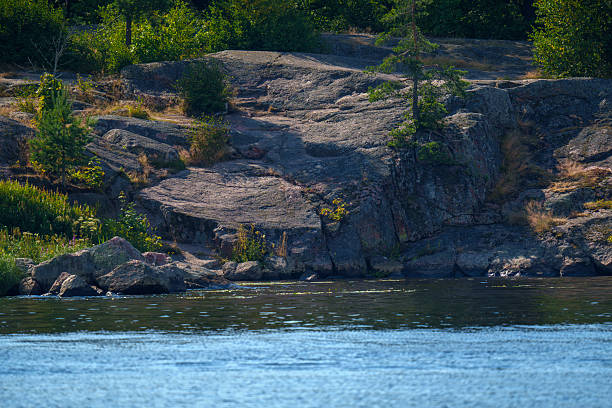 Natural rocky shoreline with layered stone formations and scattered greenery, bordering a calm lake under clear summer sunlight