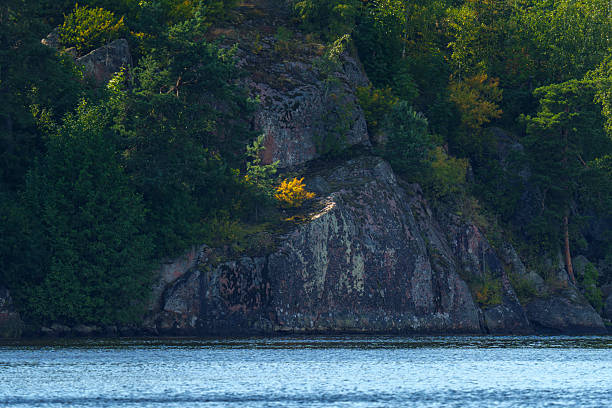 Natural rocky shoreline with layered stone formations and scattered greenery, bordering a calm lake under clear summer sunlight