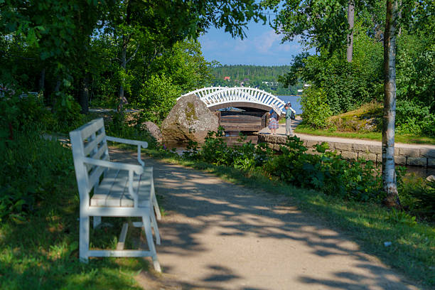 Old white wooden bench with ornate backrest stands on a sunny gravel path near green trees and wildflowers in a quiet summer park