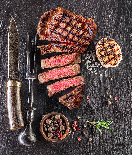 Grilled delicious ribeye steak with beef meat slices and some seasonings on black slate serving plate.
