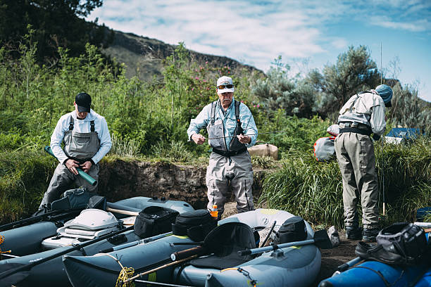 Three mature male friends prepare inflatable rafts and gear along the riverbank before beginning a multi-day fly fishing camping trip.