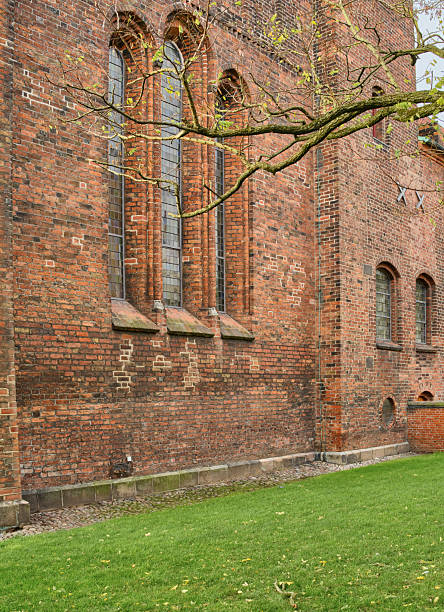 A weathered brick church exterior featuring tall arched windows and subtle stone sills, with a sprawling tree branch crossing the facade. Lush green lawn and cobblestone edge add a tranquil, historical atmosphere reminiscent of classic Danish architecture and outdoor spaces.