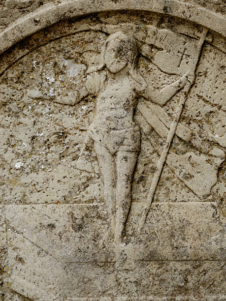 A weathered stone relief depicts a winged figure standing upright, holding a long staff with a cross at the top. The carving shows wear from time, with moss and erosion adding texture. The image evokes medieval artistry, sacred symbolism, and architectural heritage, suggesting a historic setting, possibly in Denmark.