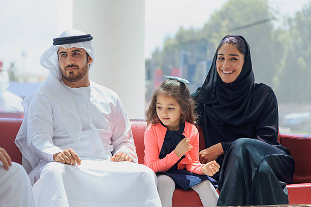 Dubai, United Arab Emirates: A traditional three generation Middle Eastern family including mother, father, grandfather and young daughter wearing traditional Arabic clothing sit together on red lounge chairs in a bright modern indoor waiting area. The family is smiling, talking and spending quality time while waiting, captured from an elevated aerial view through textured glass creating layered reflections and graphic shadows. The scene features contemporary architecture, large glass walls, natural daylight, indoor plants and a calm atmosphere suitable for shopping mall, airport terminal, hospital lobby or public building waiting area, representing family bonding, lifestyle and modern Middle Eastern culture