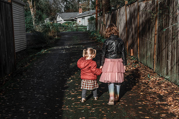 Sisters hold hands and walk down the driveway. There is a streak of sunlight and the image is faceless from behind. The toddler wears a read puffer jacket, pigtails, and plaid skirt. Her big sister wears a black leather jacket with a girly pink skirt and multi color rainbows. They are in the Pacific Northwest.