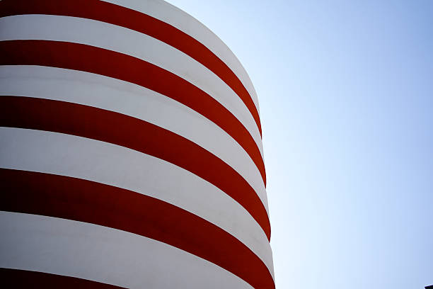 A striking architectural detail featuring a curved building facade painted with bold horizontal red and white stripes, set against a clear blue sky. The clean lines and repetitive pattern create a strong visual rhythm, evoking themes of modern design, minimalism, and urban art. This structure, likely a parking garage or public facility, showcases how functional infrastructure can be transformed into a visually compelling landmark. The contrast between the vibrant colors and the simplicity of the form highlights the beauty of geometric abstraction in contemporary architecture.