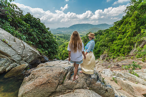 Two people holding hands on rocky viewpoint at Na Muang Waterfall 2 in Koh Samui, overlooking green valley and tropical forest during outdoor travel. High quality photo